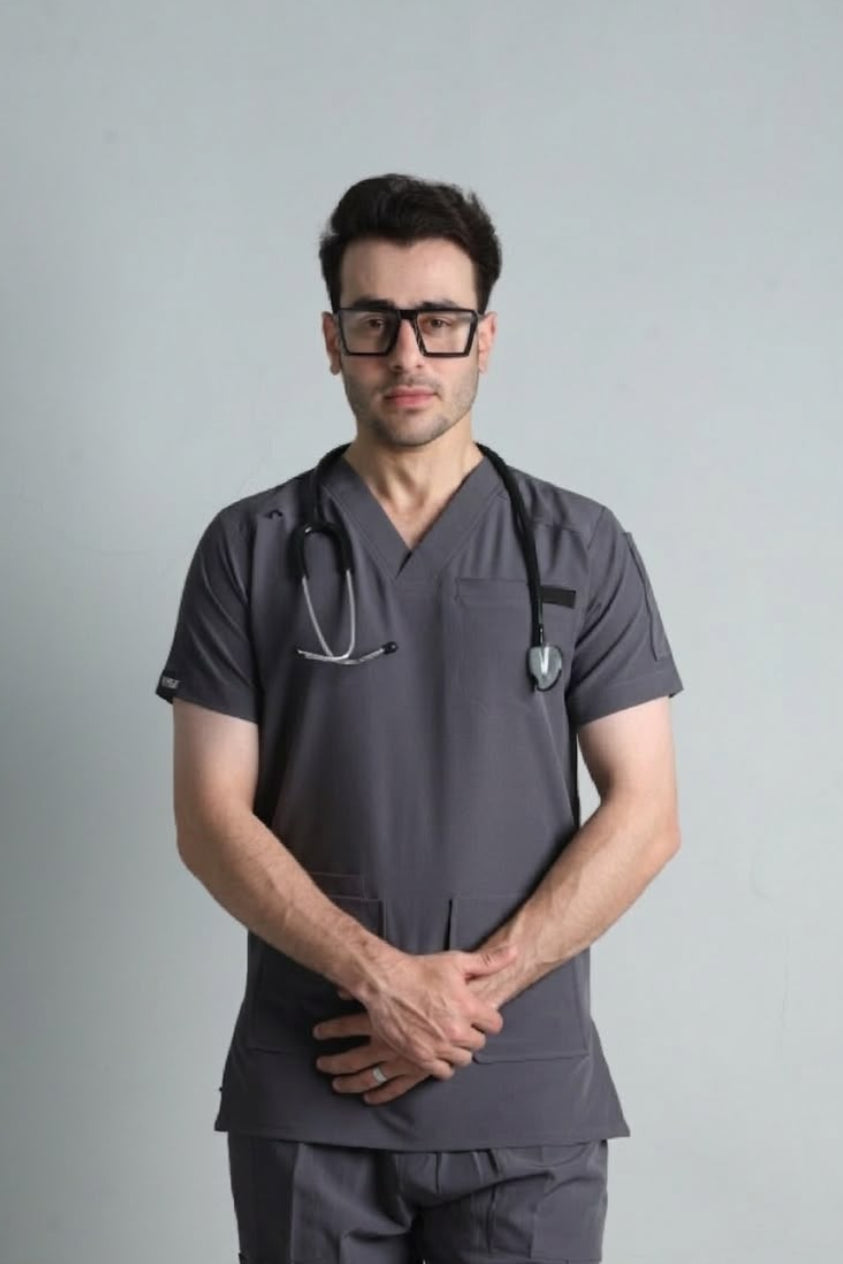 A man wearing dark gray medical scrubs and black-framed glasses, with a stethoscope around his neck, standing against a light gray studio background.