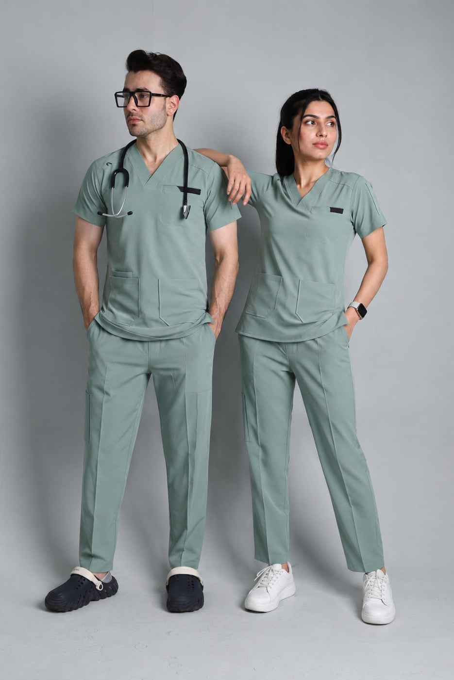 A man and a woman modeling matching sage green medical scrubs sets (short-sleeve shirts and pants) against a solid gray studio background. The man wears a stethoscope around his neck and dark loafers, while the woman wears white sneakers.