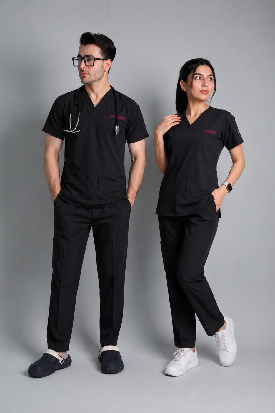 Two models wearing matching black medical scrubs sets, one male and one female, standing against a solid gray studio background. The male model wears a short-sleeve scrub top with a stethoscope and pocket, paired with black pants and dark slippers.