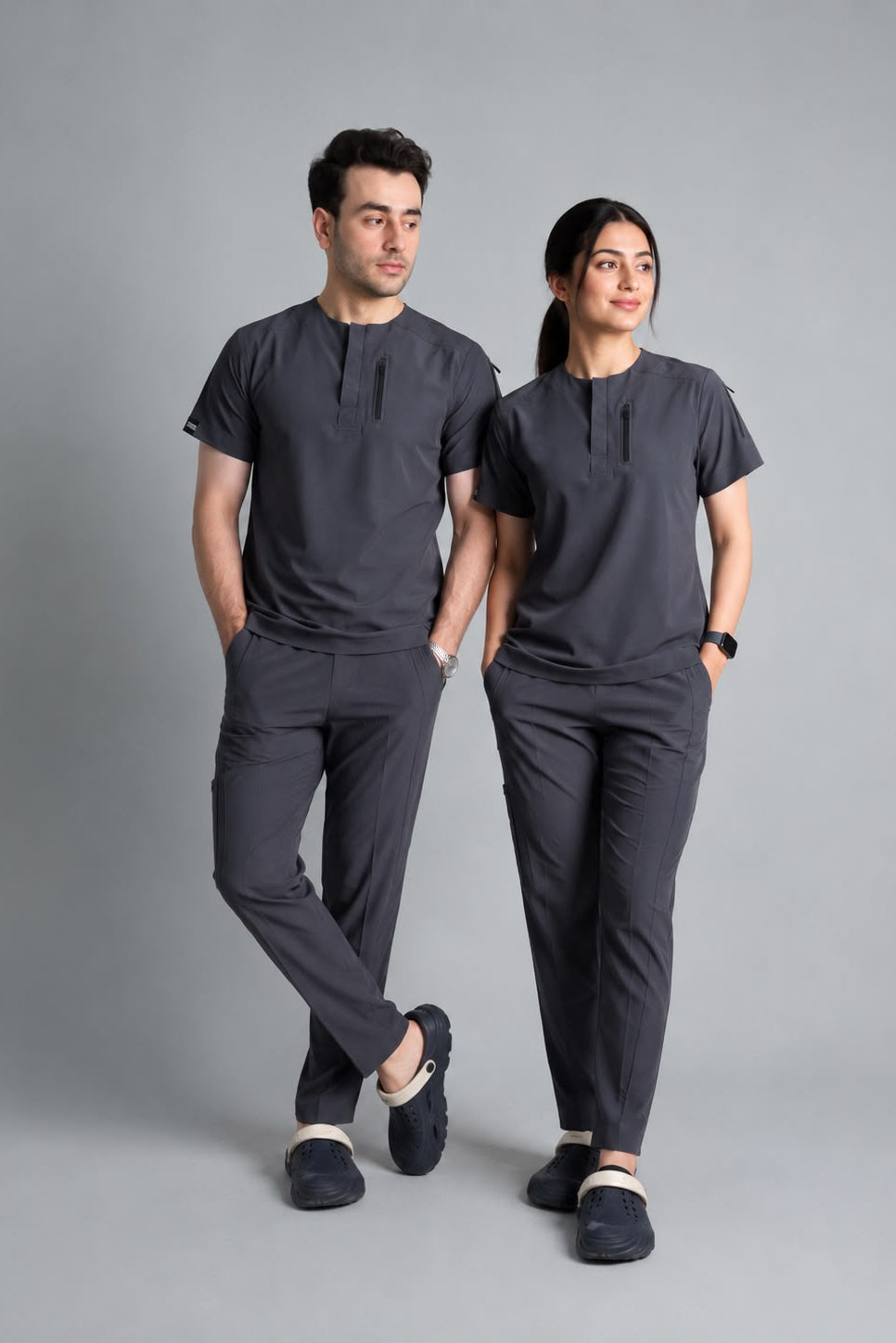 A man and a woman modeling matching dark gray scrub sets consisting of short-sleeve button-front scrub tops and scrub pants, standing against a solid gray studio background.