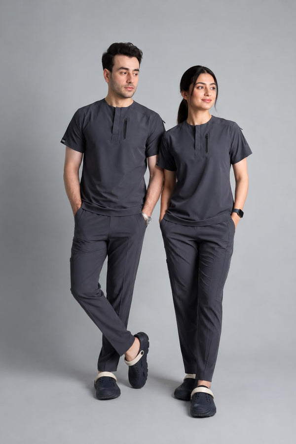 A man and a woman modeling matching dark gray scrub sets consisting of short-sleeve button-front scrub tops and scrub pants, standing against a solid gray studio background.