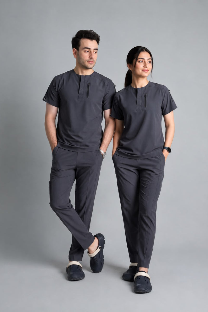A man and a woman modeling matching dark gray scrub sets consisting of short-sleeve button-front scrub tops and scrub pants, standing against a solid gray studio background.